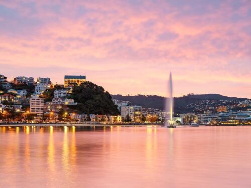 Sunset over a lakeside city with illuminated buildings and a tall fountain shooting water into the air, reflections on calm water.