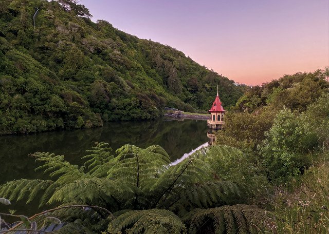 Red-roofed gazebo on a small lake island connected by a bridge, surrounded by green hills at sunset.