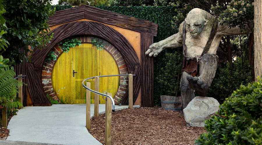 Hobbit-style round yellow door set in a wooden arch, with a garden path and a stone troll statue nearby to the right.