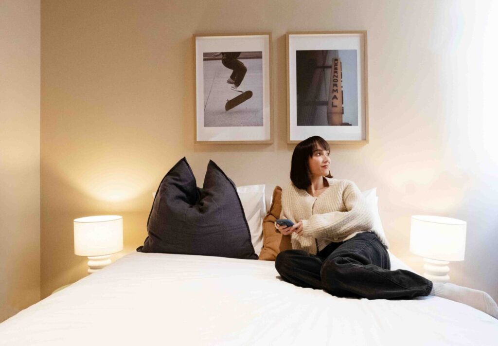 Woman lounging on a white bed in a cozy beige bedroom, holding a phone with framed wall art and warm bedside lamps nearby.