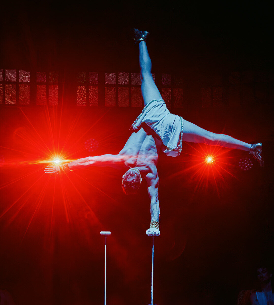 Circus performer balances on two stilts in a dramatic handstand pose, arms outstretched, wearing shorts, blue lighting with red stage lights behind.