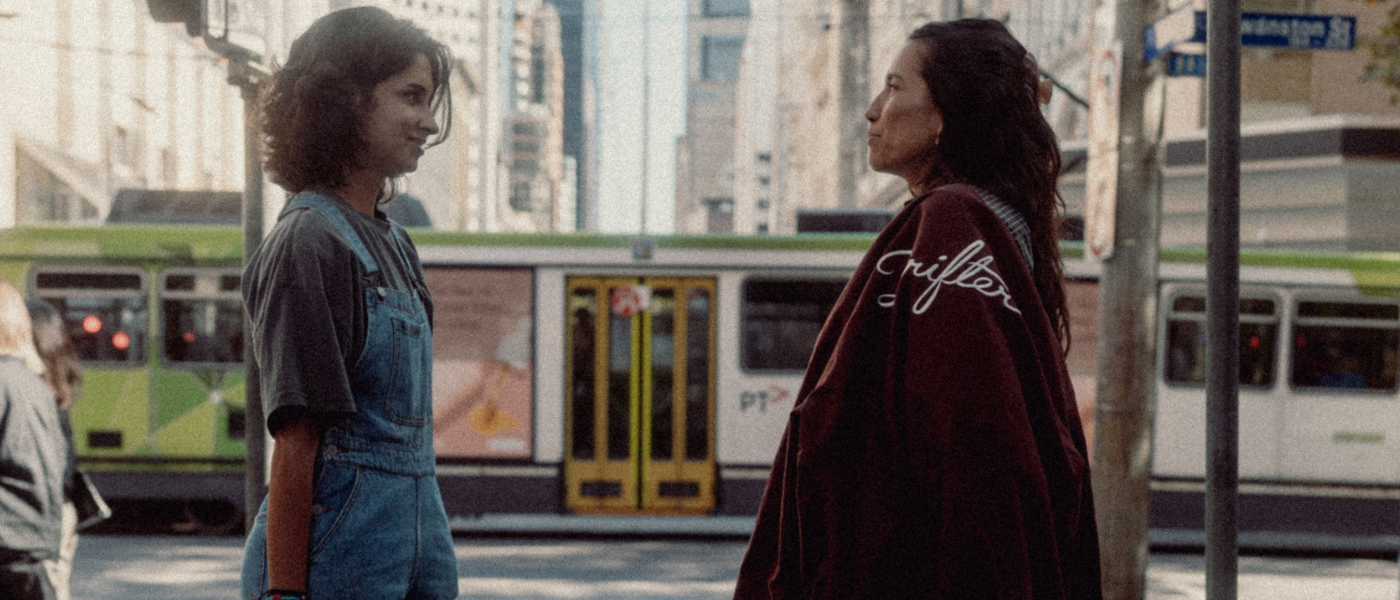 Two people stand on a city sidewalk facing each other, with a green-and-white tram in the background.