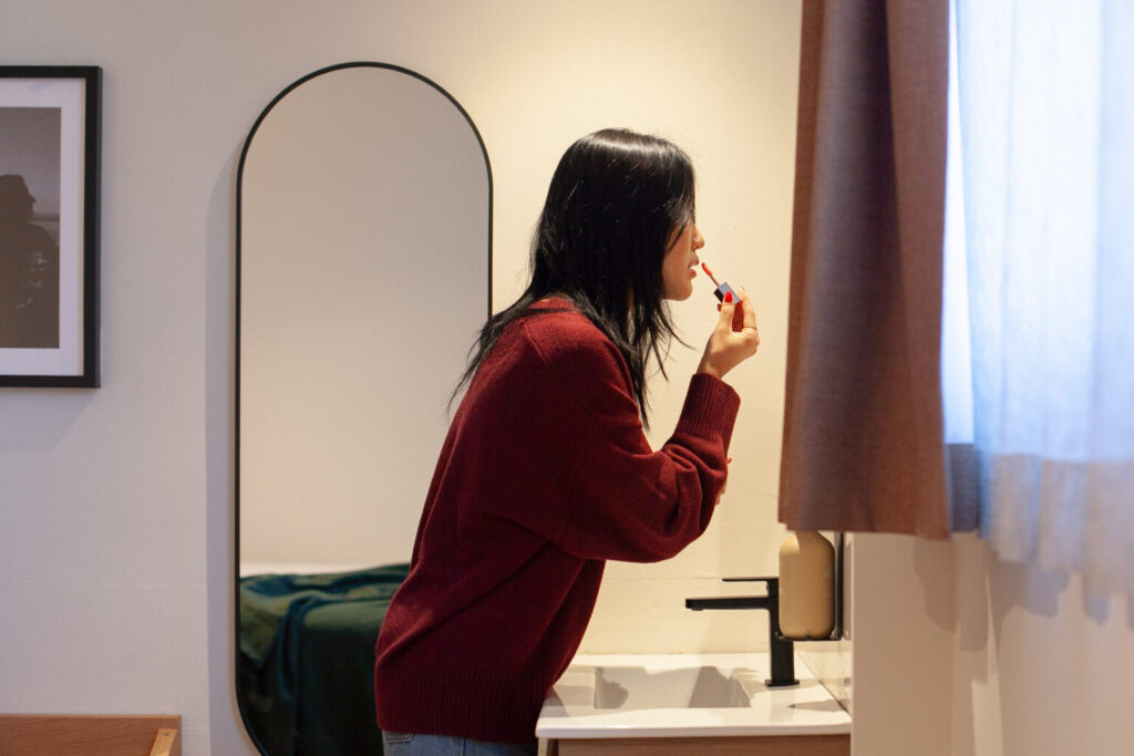 Woman in a red sweater applying lipstick at a bathroom sink, seen from the side with an oval mirror nearby.