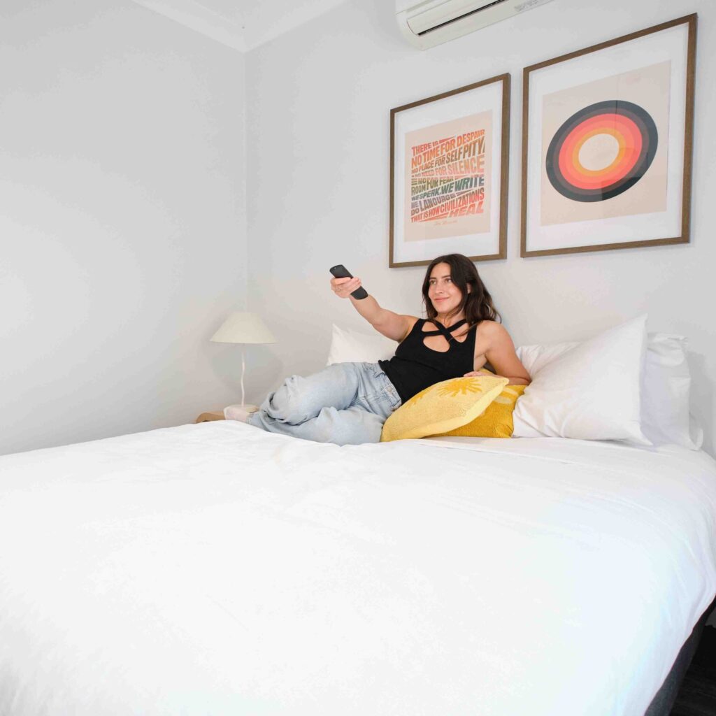 Woman lying on a bed, taking a selfie with her phone in a bright bedroom with two framed pictures above the headboard area.