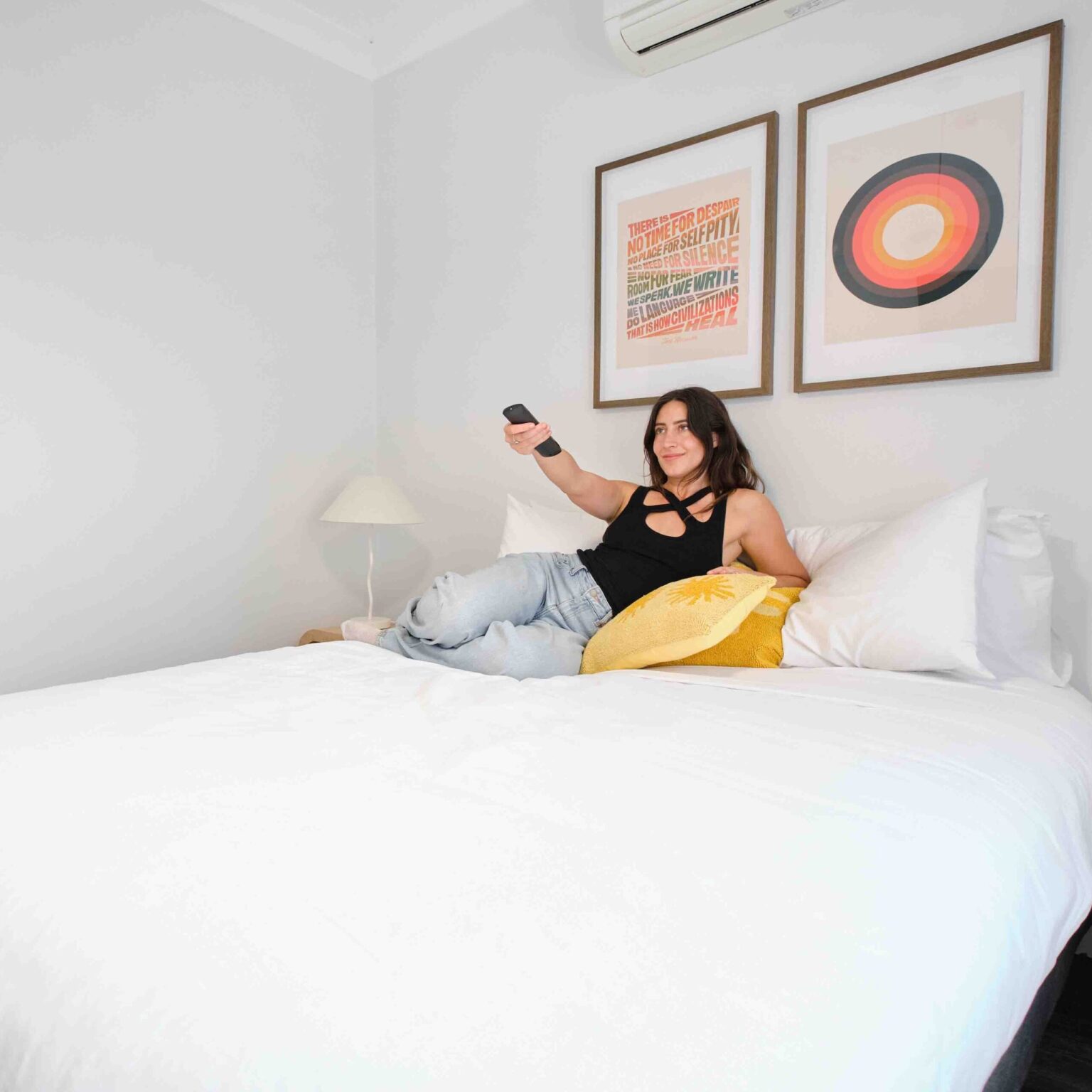 Woman lying on a white bed in a bright bedroom, taking a selfie with a smartphone; two framed abstract artworks on the wall above.