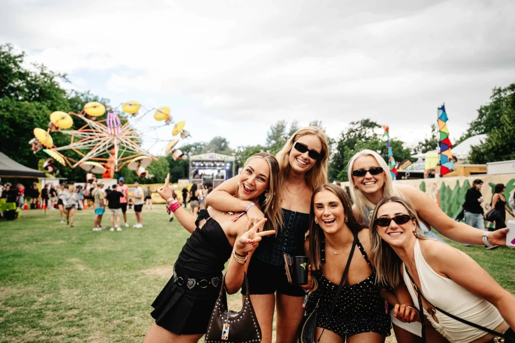 Five friends posing together at a sunny outdoor festival, with a colorful Ferris wheel in the background.