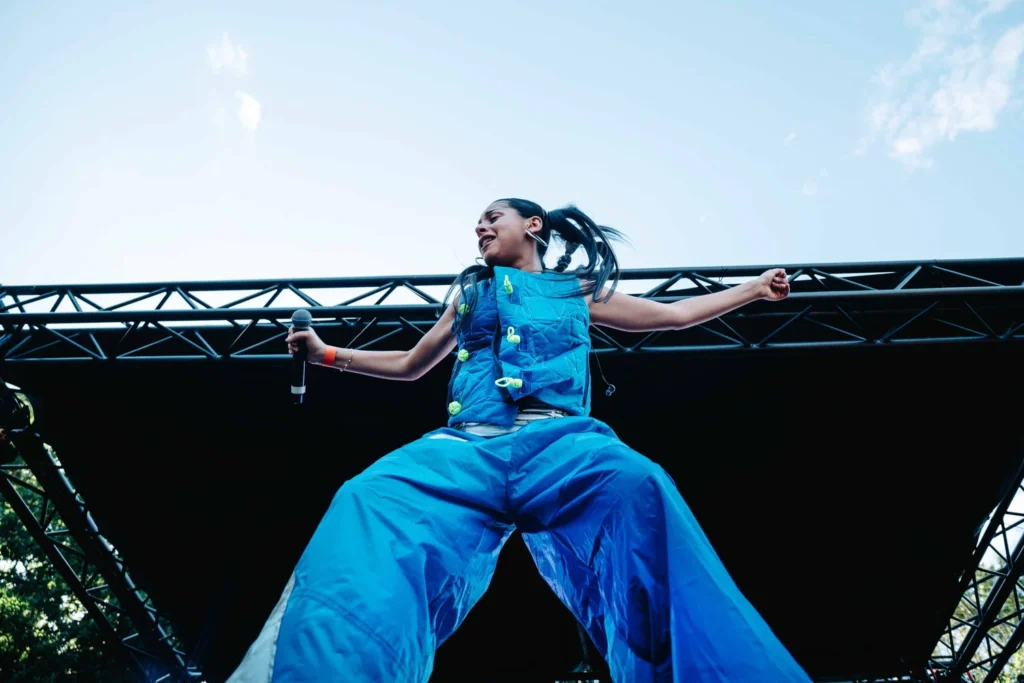 Female performer on stage in a bright blue oversized outfit, holding a microphone mid-performance.