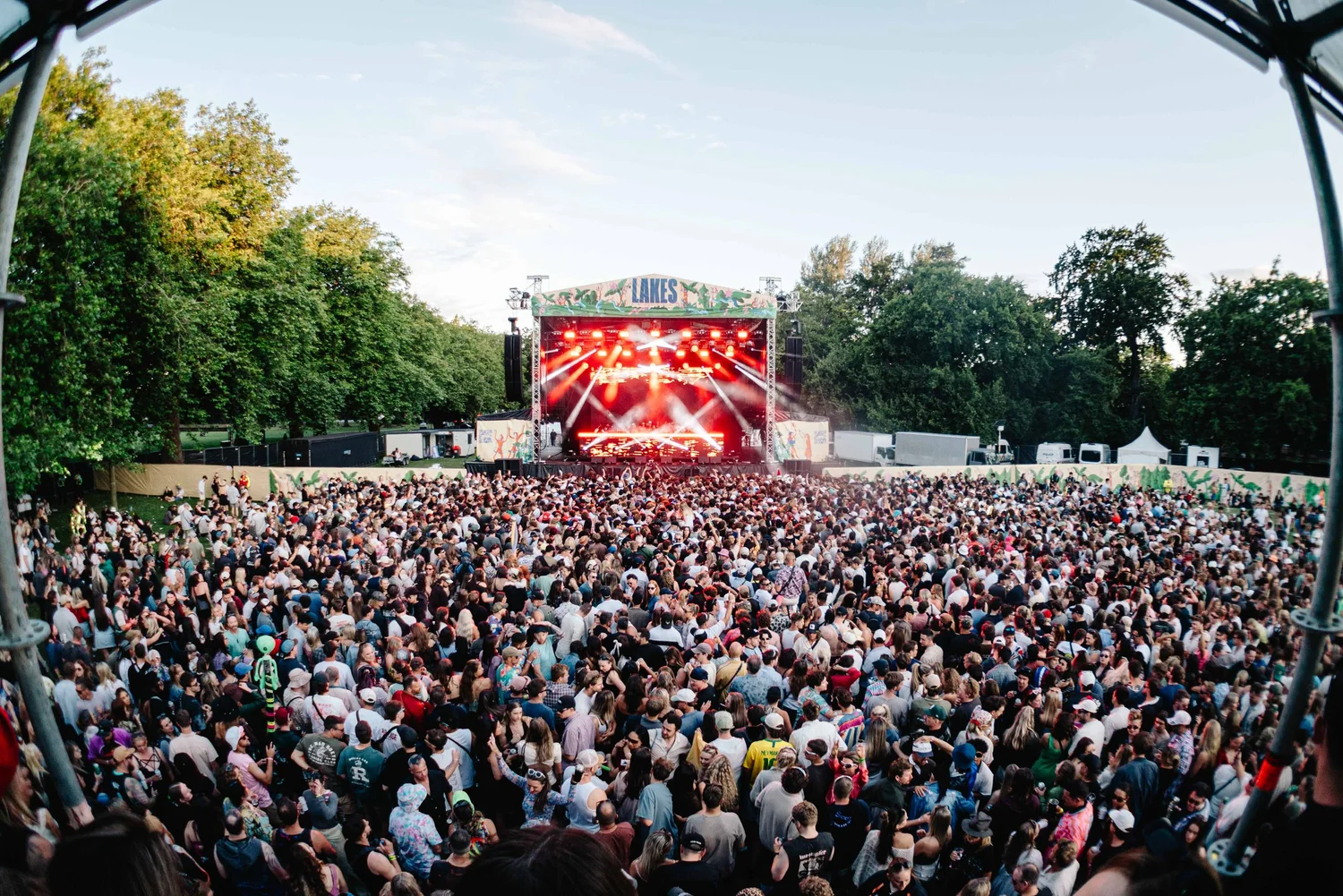 Large outdoor concert crowd at a park festival, stage with red lights and a 'LAKES' banner overhead.