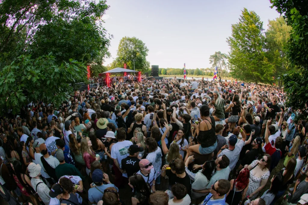 Crowd of people packed in a park festival, dancing and enjoying a stage with red lighting under a canopy of trees.