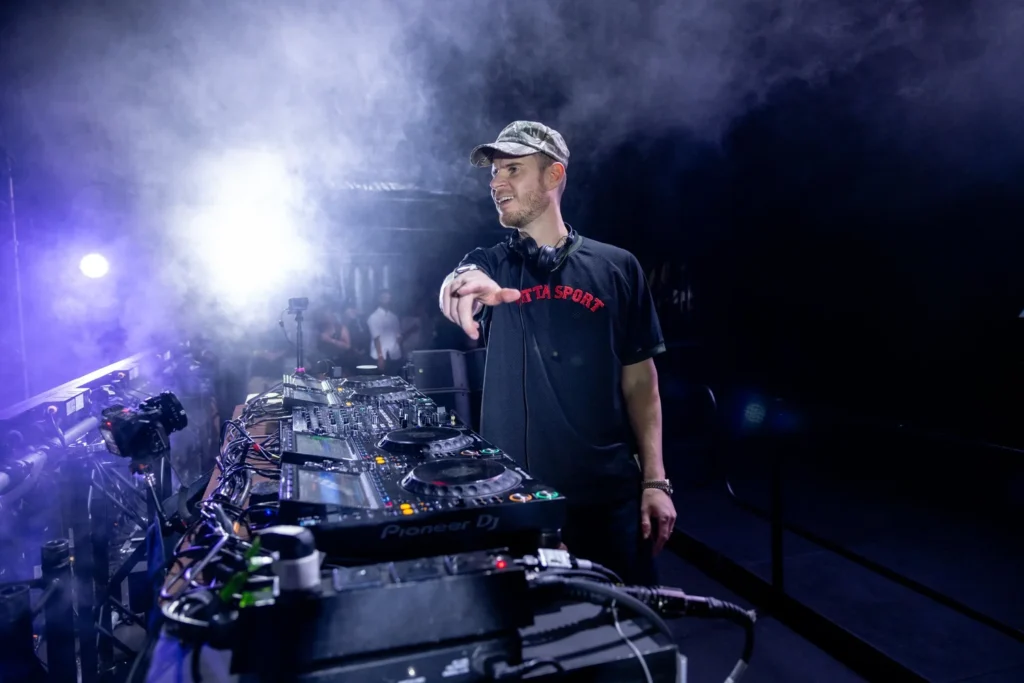 DJ wearing cap and black T-shirt behind a Pioneer DJ setup on a foggy, lit stage, pointing toward the crowd with headphones around neck