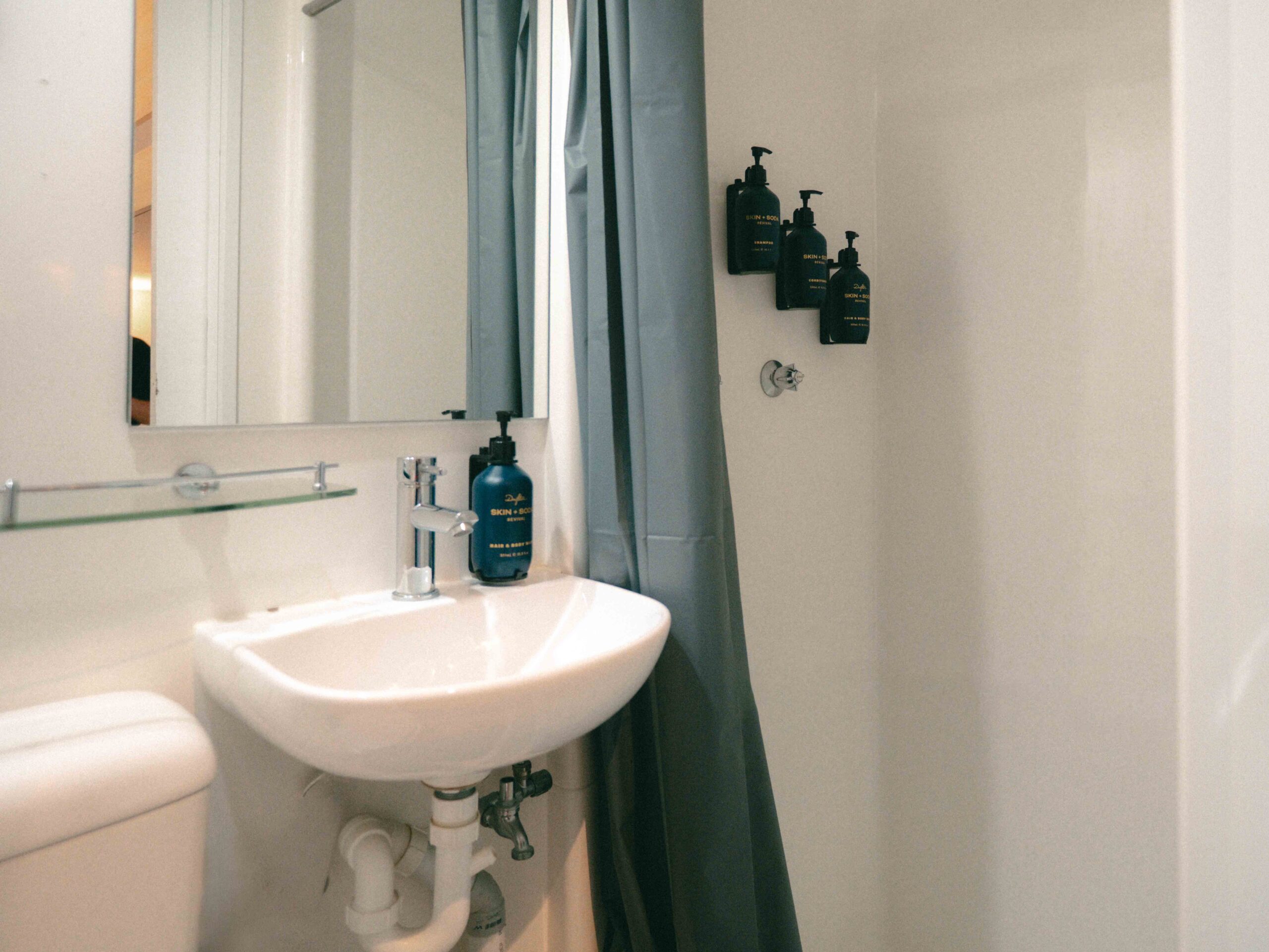 Small bathroom scene with white pedestal sink, exposed pipes beneath, and a blue bottle on the sink rim; shower curtain to the right and wall-mounted dark bottles.